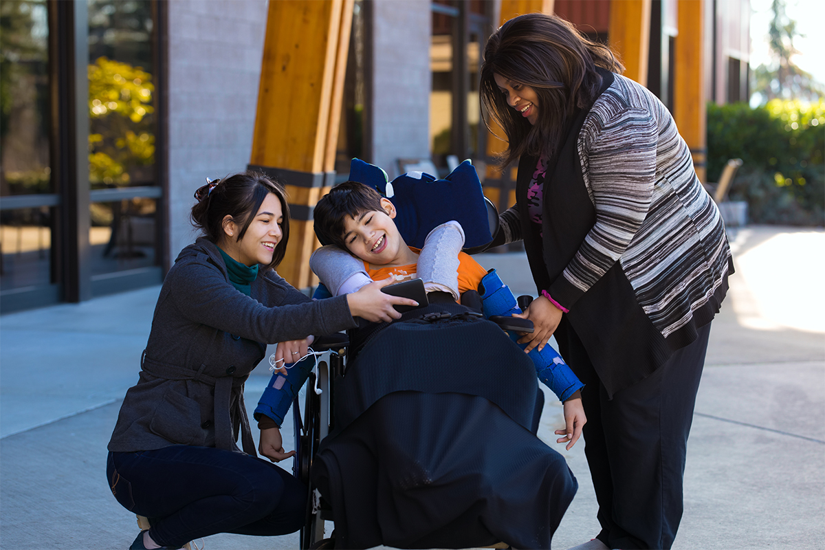 A SOCIL care team helping a smiling young person who uses a wheelchair, outdoors