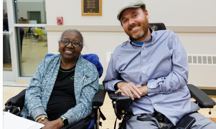 Two SOCIL community members who use wheelchairs sitting together and smiling in a community room.