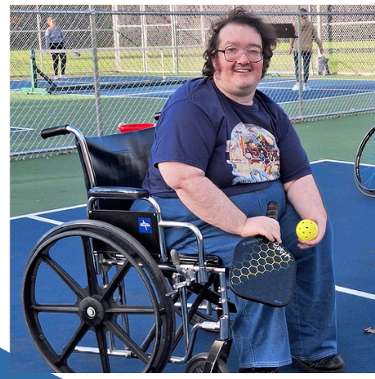 A smiling SOCIL participant in a wheelchair holding a pickleball paddle and ball on an outdoor court.