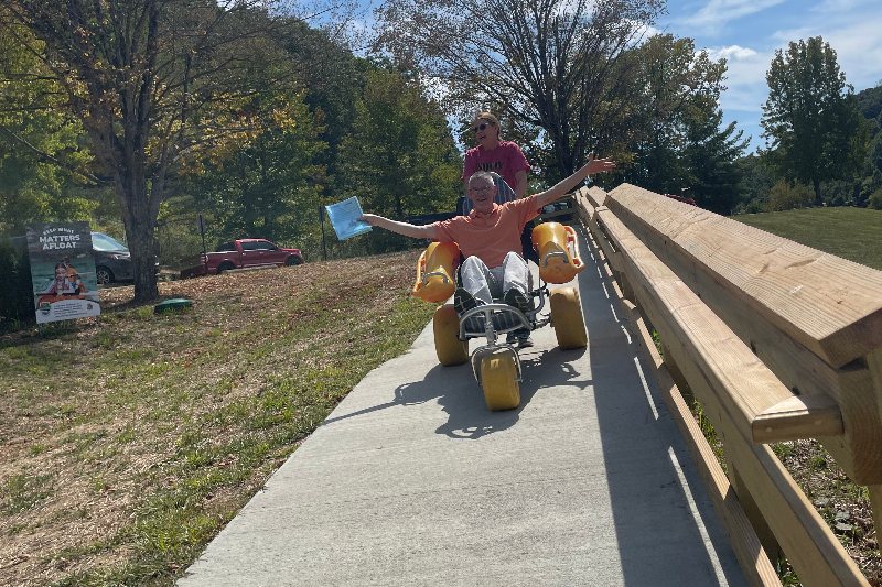 A smiling participant with arms outstretched rides a beach wheelchair down a ramp toward the lake