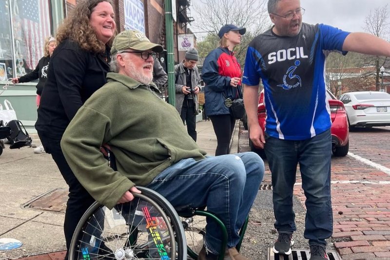 A man in a sports wheelchair laughs with friends and SOCIL staff on a downtown sidewalk