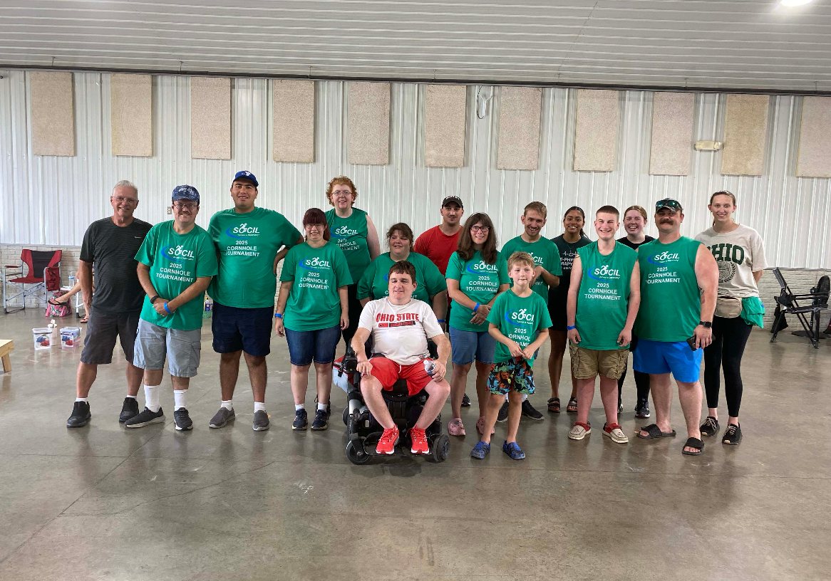 SOCIL participants and volunteers in green 2025 Cornhole Tournament shirts pose together, with a power wheelchair user in front