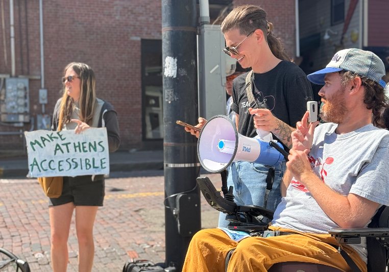 A disability advocate in a power wheelchair speaks into a megaphone beside a rally organizer, while a woman holds a Make Athens Accessible sign
