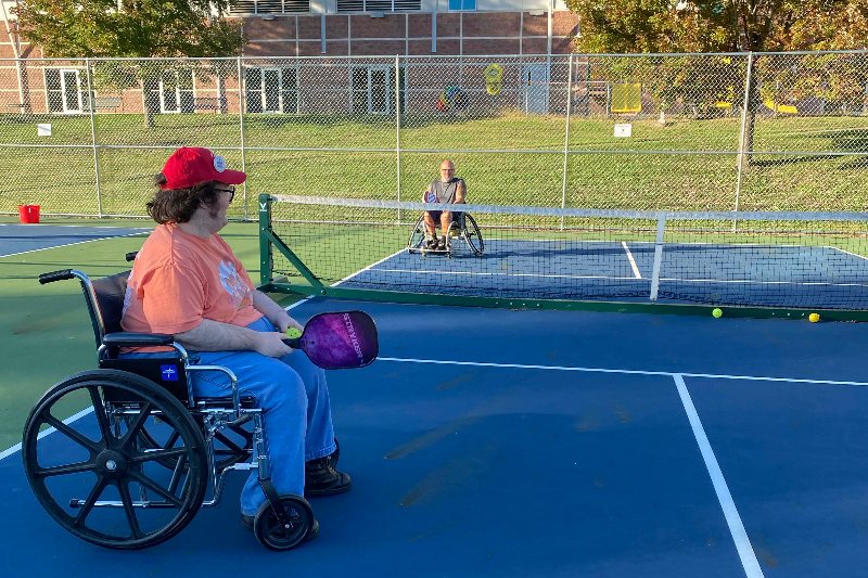 Two wheelchair users play pickleball across the net on an outdoor court